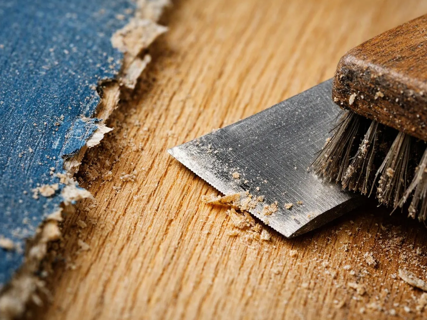 Close-up of a metal scraper beside peeling blue paint and scattered wood dust on a wooden surface