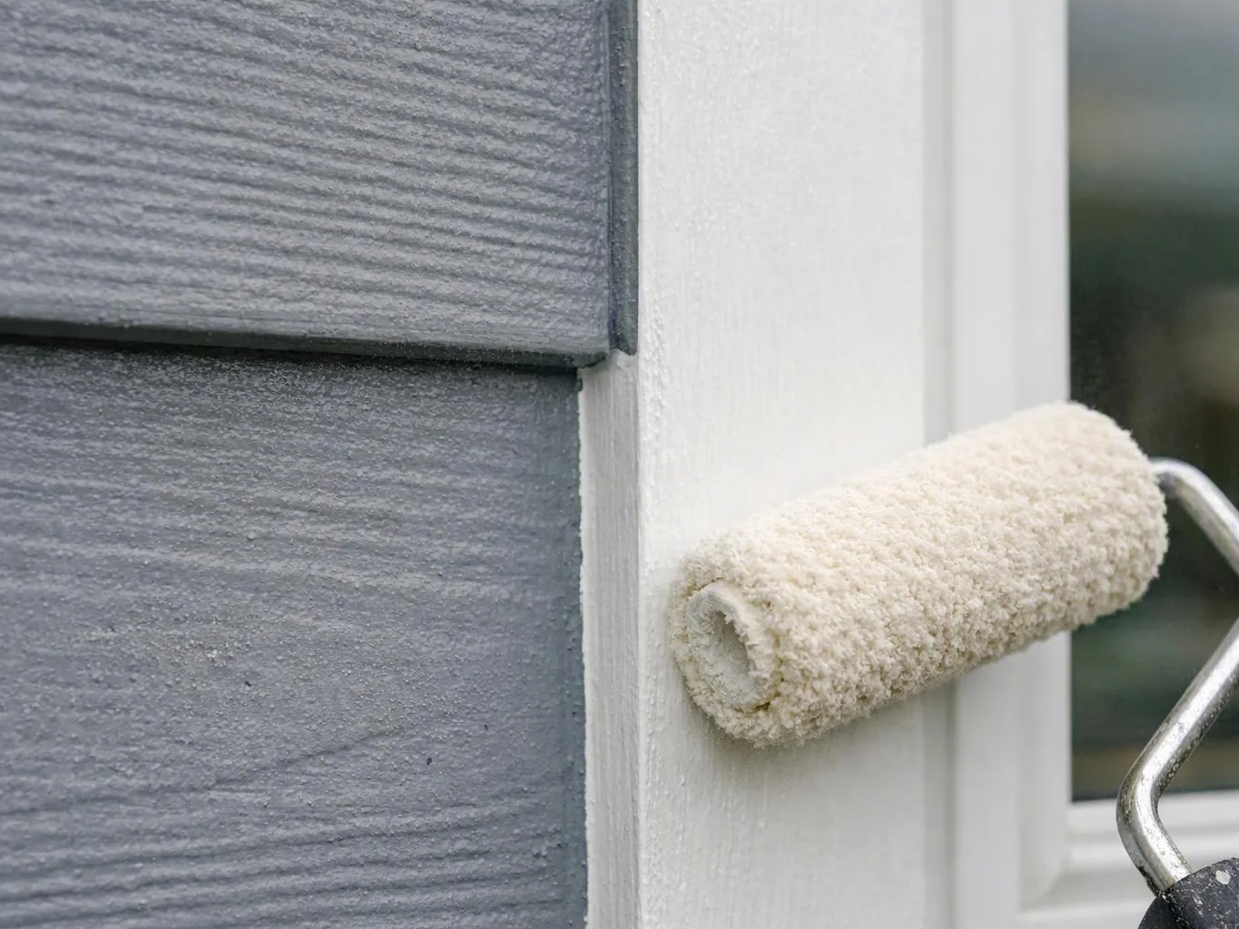 A paint roller applies white paint to trim beside gray siding and a window frame