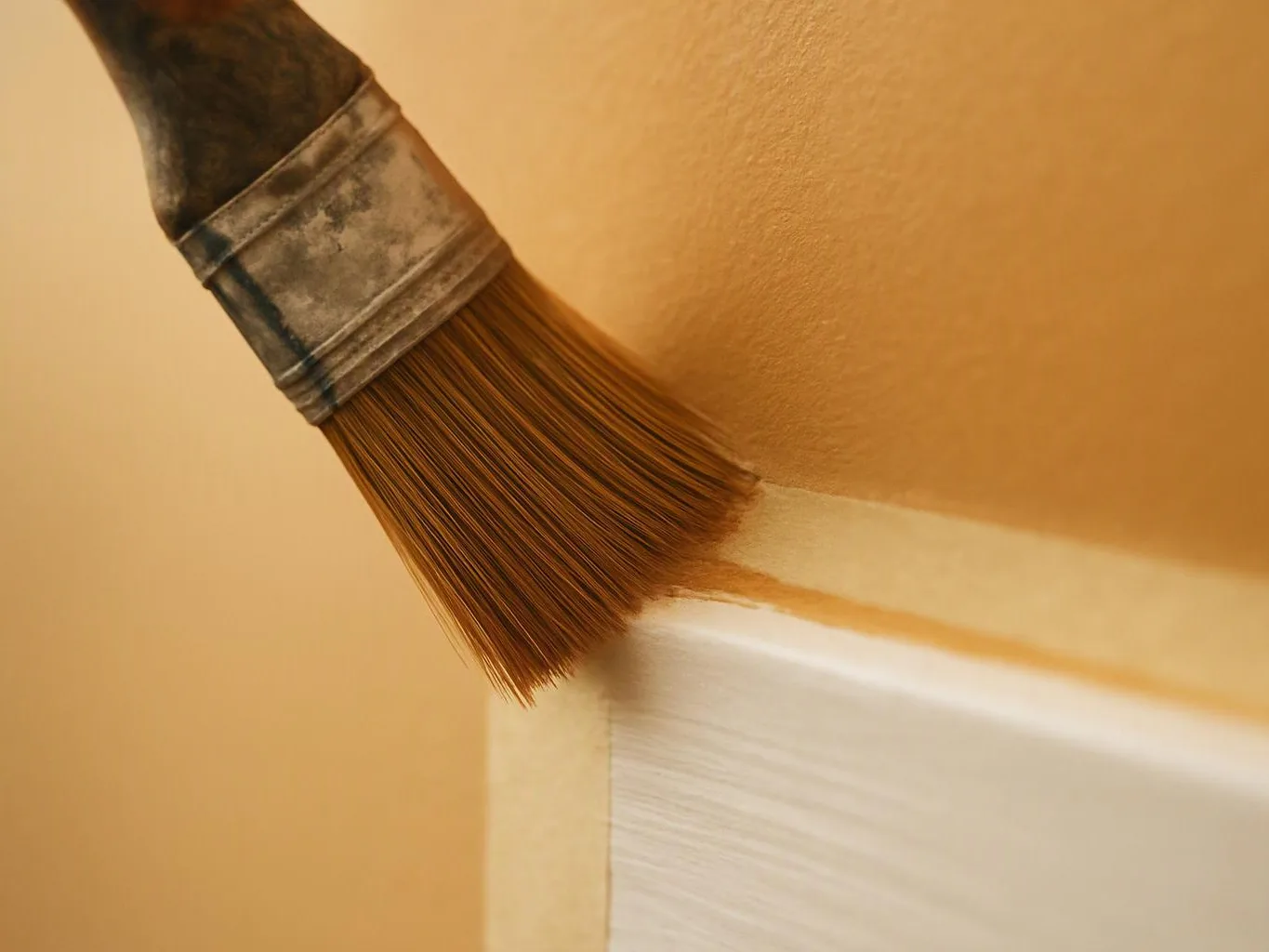 A paintbrush applies tan paint along the edge of a white trim board against a beige wall