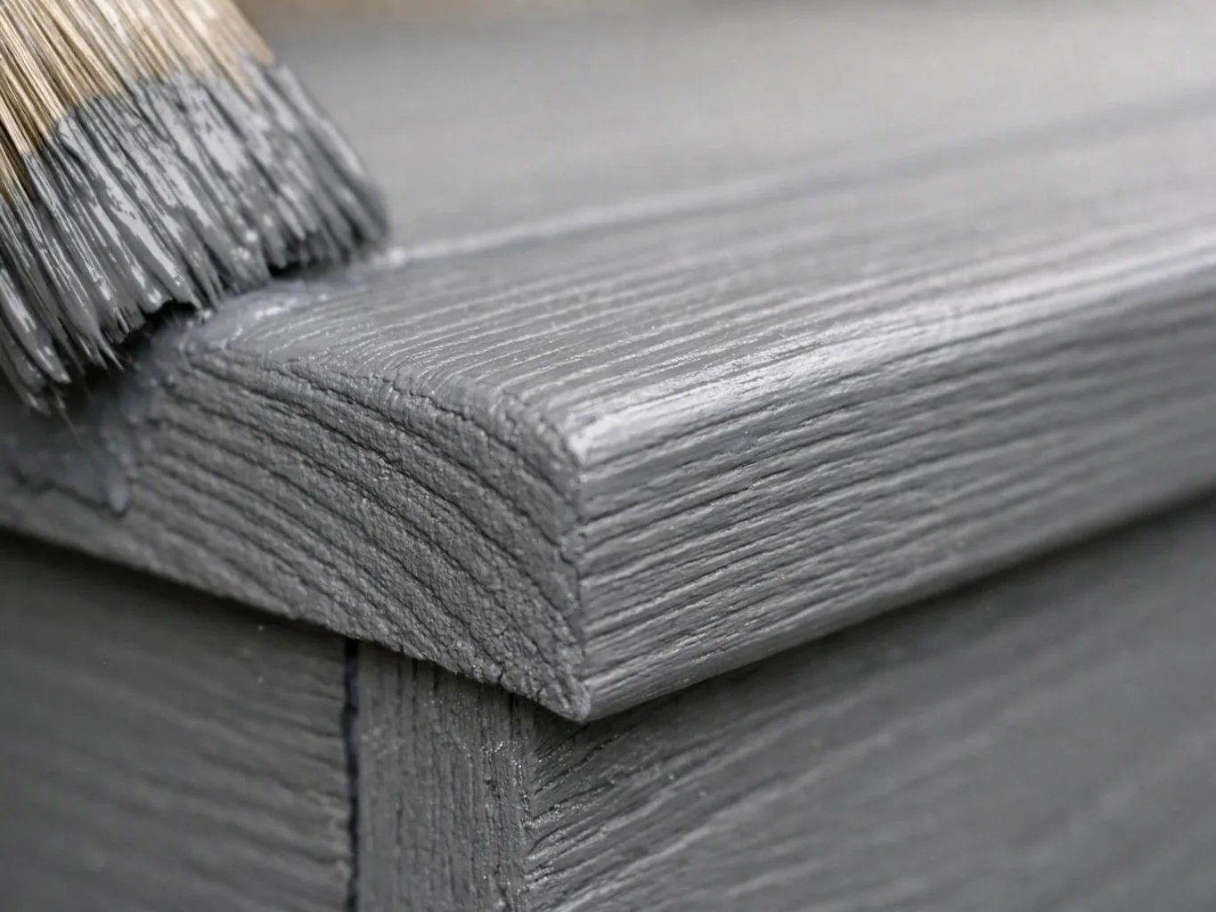 A paintbrush coats a gray wooden board, showing close-up wood grain and wet paint texture